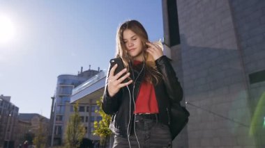 Kentsel sokak arka planda yürürken akıllı telefon kullanarak kulaklık uzun saçlı çekici genç kadın. Açık havada, yaşam tarzı