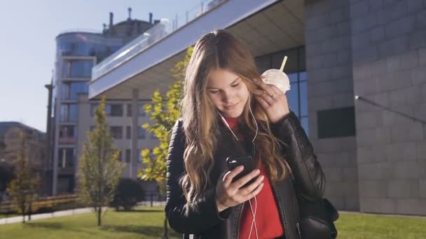 Bastante hermosa chica con el pelo largo en los auriculares utilizando el teléfono inteligente mientras escucha música y navegar por la foto interesante en la aplicación para el teléfono inteligente. Exterior, concepto urbano — Vídeo de stock