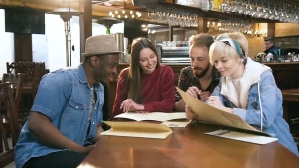 Fyra multietniska vänner väljer rätter från menyn i baren, pub. — Stockvideo