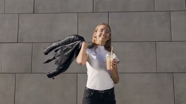 Porträtt av en ung vacker flicka i en vit T-shirt och jacka dricka milkshake genom ett rör i sommardag nära byggnad med grå-vägg bakgrund. Utomhus, livsstil — Stockvideo