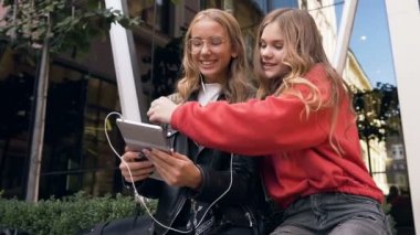 İki kadın arkadaş açık havada dijital tablet kullanıyor. Gülümseyen kız arkadaşlar tablet cihazda fotoğraflara bakarak, şehrin modern merkezi yakınında zevkle yürüyorlar. Açık havada