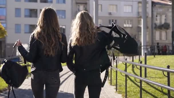 Se baksidan av två snygga unga flickvänner med långt hår och snygga ryggsäckar som går i staden och chattar glatt på varm solig dag. Utomhus, vänskap koncept — Stockvideo