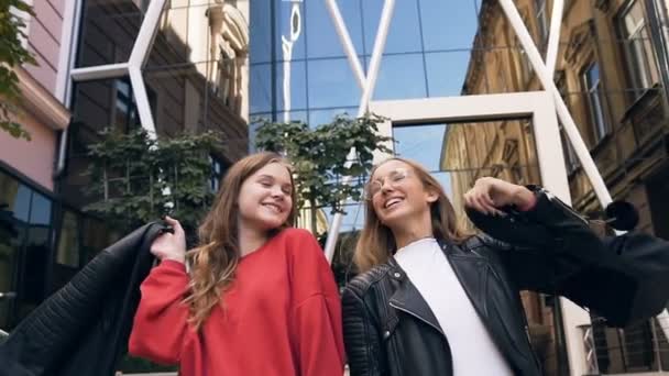 Portrait de deux gaies copines caucasiennes élégantes marchant le long de la rue près du bâtiment moderne tout en riant et en s'embrassant. En plein air, mode de vie — Video