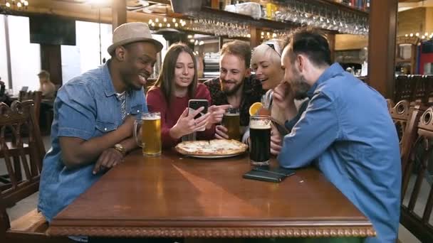 Grupo de amigos hipster felices viendo vídeo, foto en el teléfono inteligente juntos en el bar, pub . — Vídeo de stock