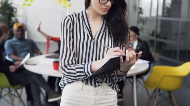 Retrato de atractiva mujer de negocios caucásica en blusa con pelo largo trabajando con documentos de papel, mirando a la cámara y sonriendo con grupo de jóvenes empresarios en el fondo — Vídeo de stock