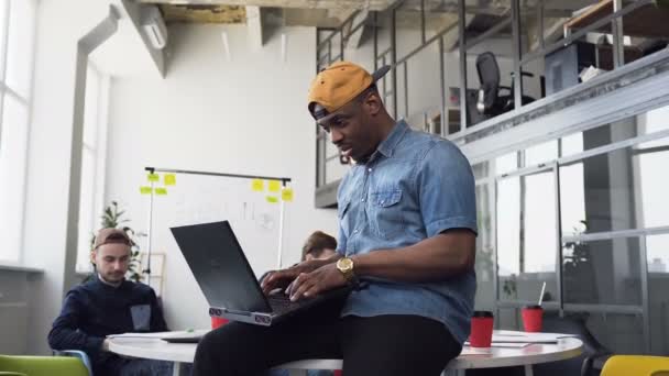 Bonito empresário afro-americano trabalhando em laptop sentado na mesa enquanto seus colegas estão sentados e trabalhando no local de trabalho em segundo plano no escritório — Vídeo de Stock