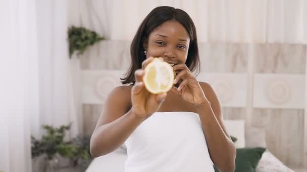Улыбающаяся молодая африканская женщина, держащая в руках киви и лимонные ломтики на светлом фоне в спа-центре. Здоровый образ жизни, здоровый уход за лицом и телом — стоковое видео
