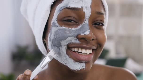 Close-up de menina afro-americana sorridente com toalha na cabeça, aplicando uma máscara de argila de limpeza no rosto, usando maquiagem escova enquanto olha para a câmera no fundo branco — Vídeo de Stock