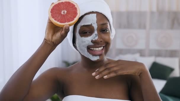 Portret van prachtige Afro-Amerikaanse vrouw in witte handdoek op hoofd Holding helft van sappige grapefruit in de voorkant van het gezicht met het toepassen van klei masker, geïsoleerd op lichte kamer achtergrond — Stockvideo