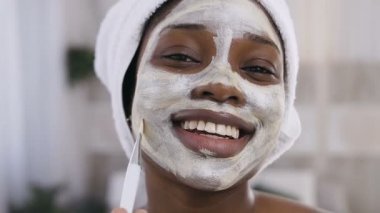 Beyaz havlu ile afro-amerikan kız yakın yüz maskesi uygulayarak ve beyaz oda arka planda gülümseyerek kafasına. Güzellik kozmetolojisi, yüz ve cilt bakım konsepti