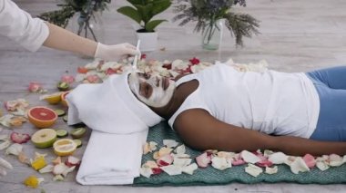 Güzel genç afro amerikan kadın kapalı gözlerle güzellik merkezinde dinleniyor kozmetolojist yüz maskesi uygularken