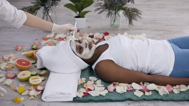 Pretty young afro american woman resting with closed eyes in beauty centre with closed eyes while cosmetologist applying facial mask — Stock Video