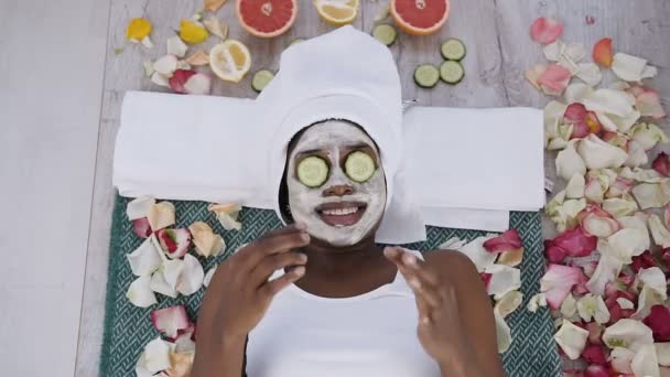 Draufsicht der schönen jungen Afrikanerin in weißem Handtuch auf dem Kopf liegend mit Gurkenstücken im Gesicht im Schönheitssalon. Schönheitsbehandlung. — Stockvideo