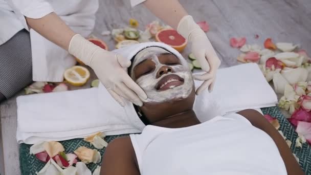 Top view of young african american woman in white towel on head lying on the couch relaxing while cosmetologist applying a cosmetic cream does face massage in beauty centre. Facial skin care. Peeling — Stock Video