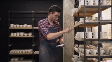 Raflardan cermics sayma ve not defterinde belirterek odaklanmış erkek çömlekçi.
