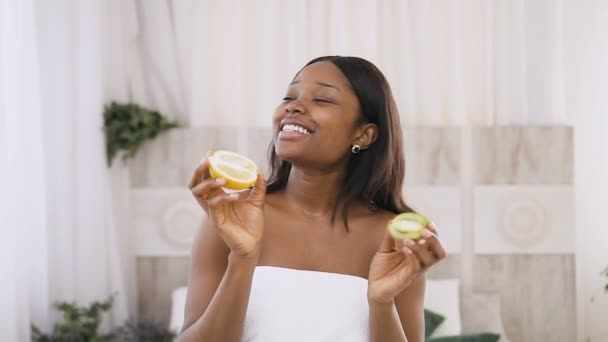 Retrato de una divertida y divertida joven afroamericana sosteniendo las mitades de kiwi y naranja en las manos sobre un fondo claro en el spa. Cuidado de la piel saludable. concepto de estilo de vida saludable — Vídeos de Stock