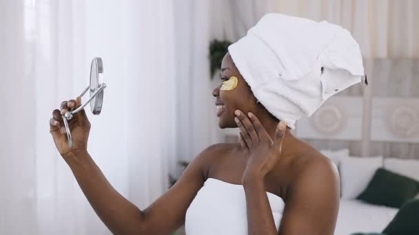 Femme afro-américaine souriante applique des taches dorées sous les yeux, a une serviette blanche sur la tête, regardant dans le miroir, dans la chambre blanche après la douche — Video