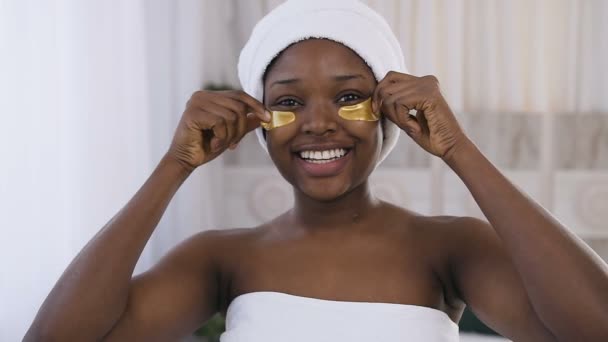 Красивая молодая африканская женщина улыбается, пока снимает медицинские золотые пятна в спальне. Здоровый образ жизни, молодежь, лица, анти-старение — стоковое видео