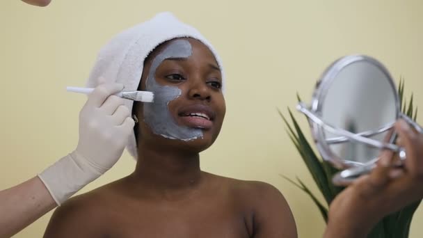 Σκοποβολή του αισθητηρίου χέρι στην εφαρμογή μάσκα στο πρόσωπο της νεαρής Αφροαμερικανικής γυναίκας στο σαλόνι σπα — Αρχείο Βίντεο