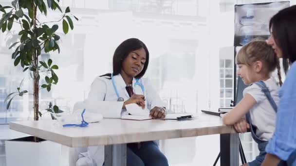Jeune médecin africain donnant la prescription à la mère mignonne avec sa fille à la clinique . — Video