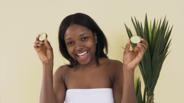 Retrato de mulher americana africana atraente removendo fatias de pepinos dos olhos e sorrindo para a câmera. Conceito de rosto saudável de cuidados com a pele — Vídeo de Stock