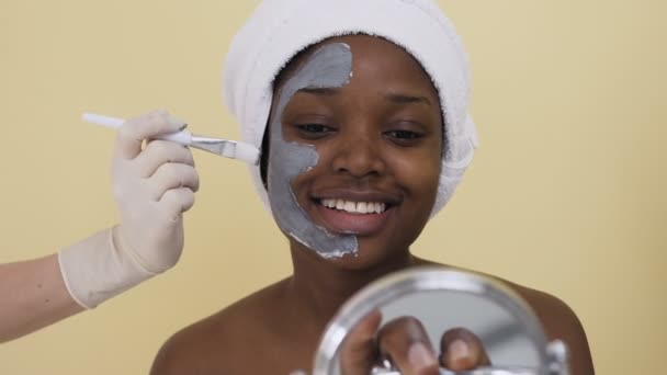 Tir de la main du cosmétologue sur l'application du masque sur le visage de la jeune femme afro-américaine dans le salon de spa — Video