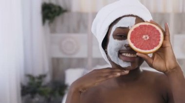Beyaz oda arka plan greyfurt yarıya tutarken baş beyaz havlu genç afro amerikan kadın yarım yüz yüz yüz kil maskesi uygulanan