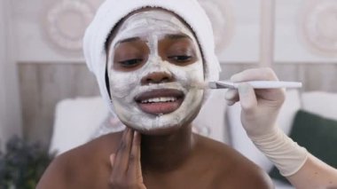 Yakından kapatın. Spa merkezinde özel fırça ile yüzüne maske uygulayan kozmetolojist el başında havlu ile Afrikalı-Amerikalı kadın