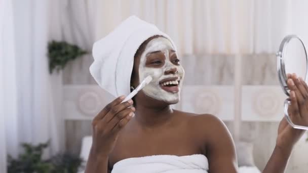 Mujer afroamericana feliz en toalla blanca en la cabeza aplicando mascarilla de yogur en la cara, mirando en el espejo. Concepto saludable de facial, centelleo y rejuvenecimiento — Vídeos de Stock