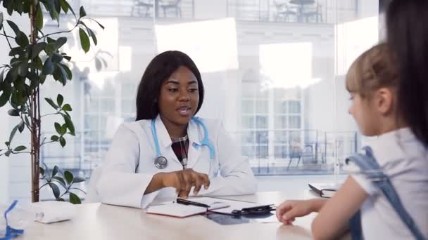 Joven médico africano dando cinco a la niña que está sentada en las rodillas de su madre . — Vídeos de Stock