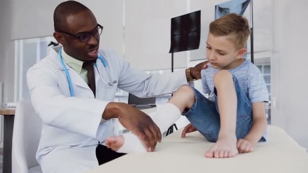 Чоловік лікар оглядає пов'язку на нозі маленького хлопчика . — стокове відео