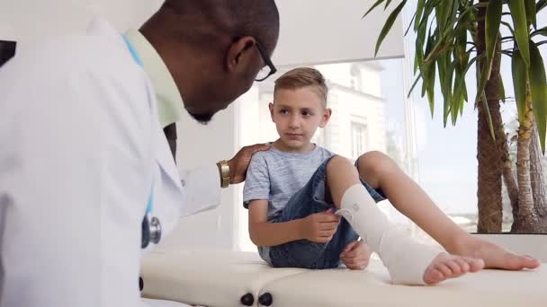 Afrikansk läkare pratar med liten patient med bandage på benet. — Stockvideo