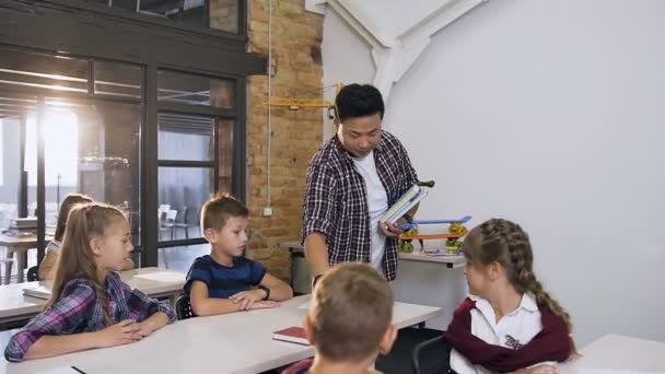 Jonge Koreaanse leraar lopen tussen tussen bureaus zet schoolboeken op het Bureau, terwijl Kaukasische leerlingen zittend aan de bureaus — Stockvideo