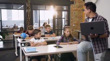 Altı ilkokul öğrencisine ders veren dizüstü bilgisayarlı Koreli erkek öğretmen. Masalarda oturan okul çocukları koreli öğretmenlerinin sorularını cevaplamak için ellerini kıyor lar.
