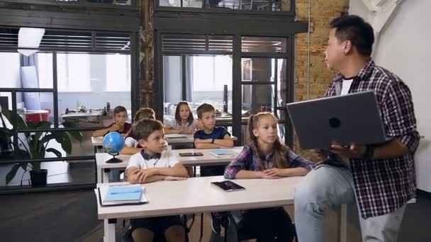 Junge koreanische männliche Lehrer sitzen am Schreibtisch mit Laptop gibt Unterricht für sechs Grundschüler. Schüler sitzen am Schreibtisch und hören zu und beantworten Fragen ihres koreanischen Lehrers — Stockvideo