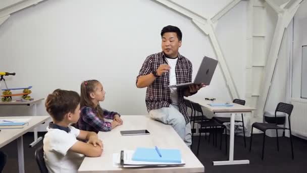 Profesor coreano sentado en el escritorio con portátil en la mano dando lección para los alumnos. Los escolares escuchan atentamente y hacen preguntas importantes a su maestro rorense — Vídeo de stock