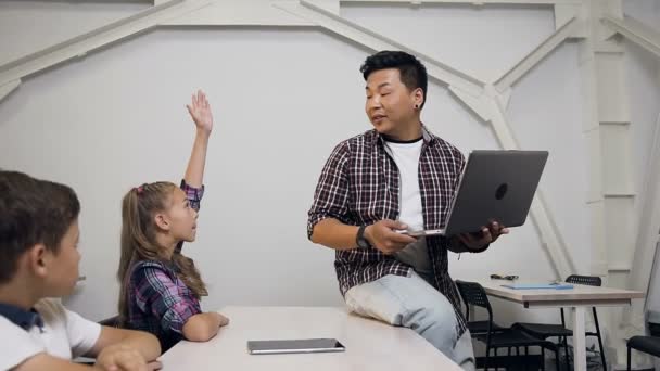 Прекрасні кавказькі школярки сидять за столом разом з однокласником, піднімаючи руку і задавати питання корейського вчителя під час уроку в школі — стокове відео