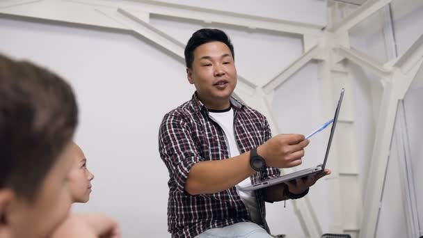 Retrato de professor coreano sentado na mesa com laptop na mão dando aula para os alunos. Alunos da escola primária ouvindo cuidadosamente palestra e fazer perguntas importantes seu professor — Vídeo de Stock