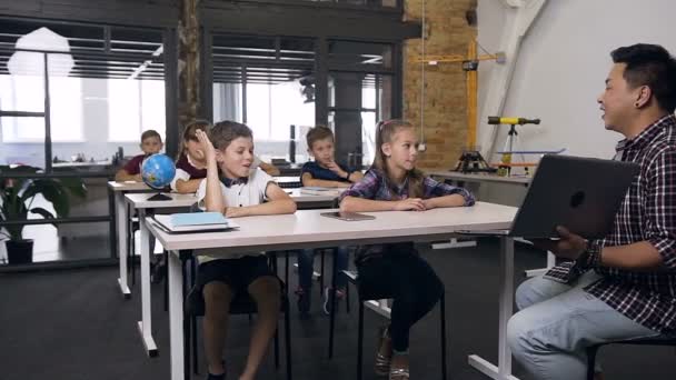 Söt kaukasiska pojke sitter bakom skrivbord tillsammans med klasskamrat, höja hand och ställa frågor till Koreanska lärare under lektionen i skolan — Stockvideo