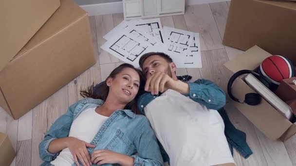 Pareja feliz tumbada en el piso en el nuevo apartamento hablando y mirando a su alrededor durante el proceso de movimiento. Relación y concepto de casa . — Vídeos de Stock