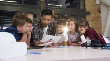 Koreli erkek öğretmenlerinokulda elektronik tasarımcı çalıştırmak için tablet bilgisayar cihazını kullanırken, farklı etnik kökenlerden gülümseyen altı meraklı okul çocuğundan oluşan grubun vurulması