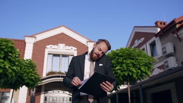 集中した若いあごひげのオフィスワーカーは、彼の携帯電話で話し、素敵な家の近くで屋外で文書を見て — ストック動画