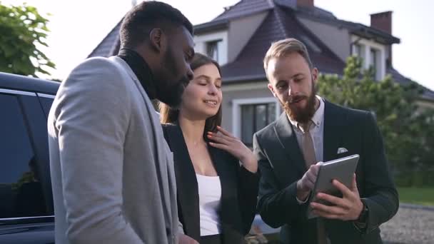 Yakışıklı sakallı araba satıcısı tablet kullanarak, satın alınan otomobil müşterilerine özelliklerini gösteren — Stok video