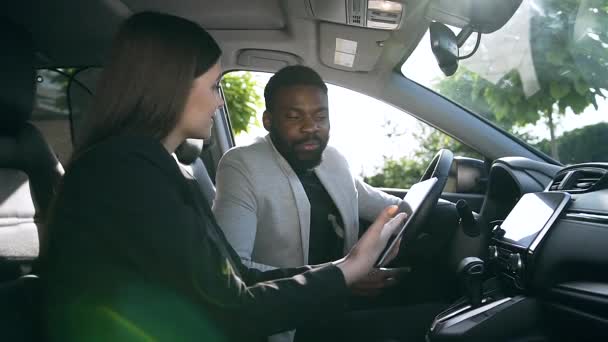 Mooie vrouw met lang haar praten met elegante Afro-Amerikaanse man, zittend in de voor stoelen van zijn luxe auto — Stockvideo