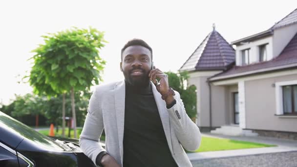 Веселий молодий афроамериканець людина в стильному одязі стояв біля чорного автомобіля і говорити по телефону з щасливою посмішкою — стокове відео