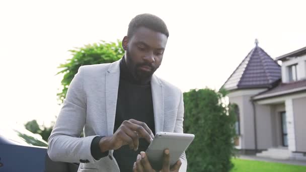 Grave barbudo hombre afroamericano de pie cerca de coche negro en el patio de la casa acogedora y el uso de su tableta — Vídeo de stock