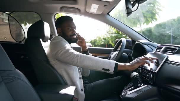 Elegante hombre de negocios afroamericano bullicioso sentado en el asiento del conductor, hablando por teléfono y sintonizando el coche en la barra de tareas — Vídeo de stock