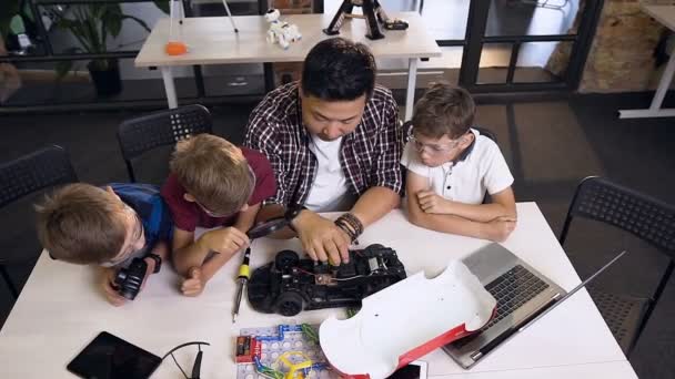 Visão superior do professor com crianças pequenas tentando reparar placa de computador no protótipo de uma máquina elétrica na escola de engenharia — Vídeo de Stock