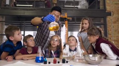 Bilim adamı çocuklara laboratuvar sınıfında kimyasal reaksiyon deneylerini nasıl yapacaklarını gösteriyor. Gözlük şişesinde kimyasal reaksiyon turuncu renk. Bilim, kimya ve bilim adamları