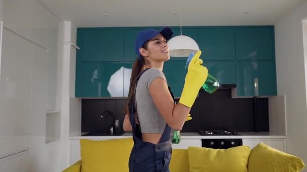 Tetszetős 30s lány egységes gazdaságban két palack mosószerek, permetezés, és táncol a konyhában — Stock videók